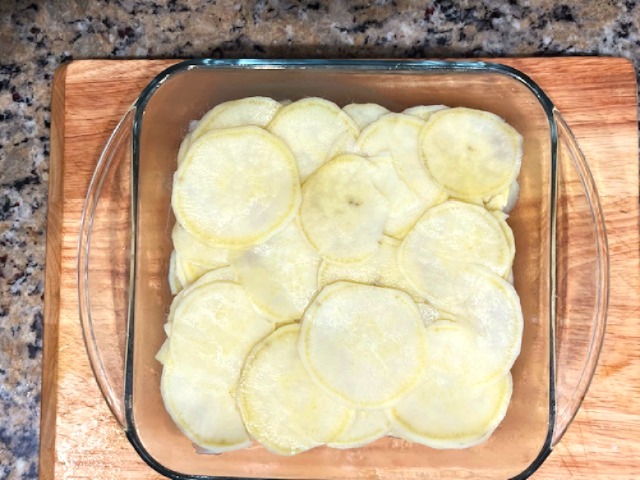 Parboiled sweet potato slices layered in a glass baking dish, ready to be topped with shrimp and coconut milk sauce for an AIP Reintroduction Garlic Shrimp and Sweet Potato Gratin.