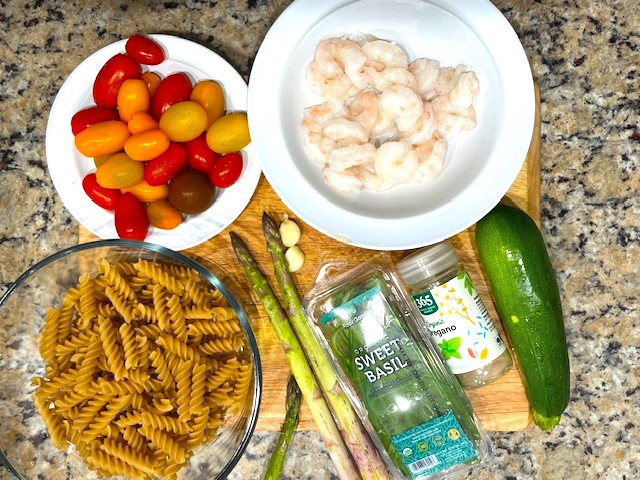 Fresh ingredients for Shrimp and Veggie Pasta arranged on a cutting board, including zucchini, asparagus, cherry tomatoes, and shrimp. A fresh and healthy gluten-free, dairy-free, and AIP Reintroduction meal.