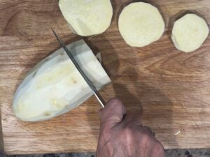 Hand slicing a peeled white sweet potato into thick rounds on a wooden cutting board for Deviled No Eggs Potatoes.