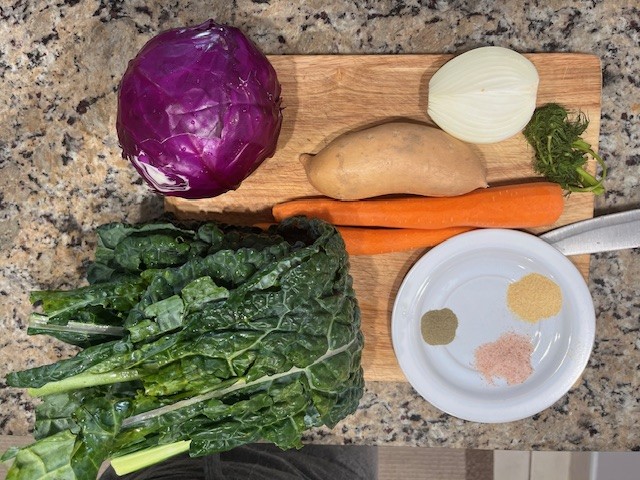Fresh ingredients for Sweet Potato Cabbage Stew including red cabbage, kale, carrots, sweet potato, onion, dill, and spices on a wooden board.