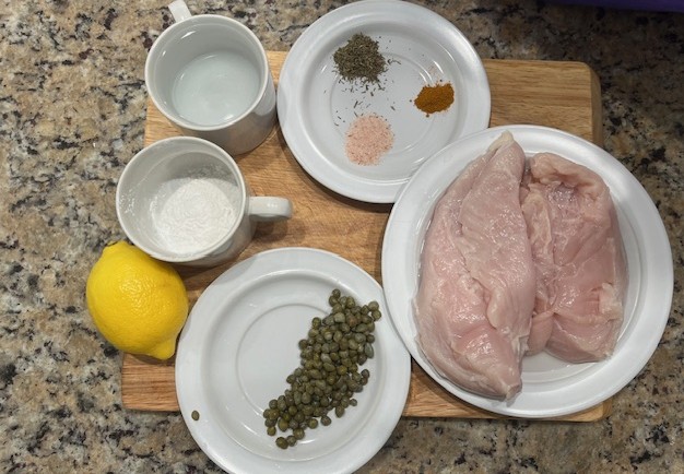 Healthiest AIP chicken piccata ingredients including chicken breast, lemon, capers, tapioca flour, sea salt, turmeric, herbs, and bone broth on a wooden board for a gluten free and dairy free recipe.